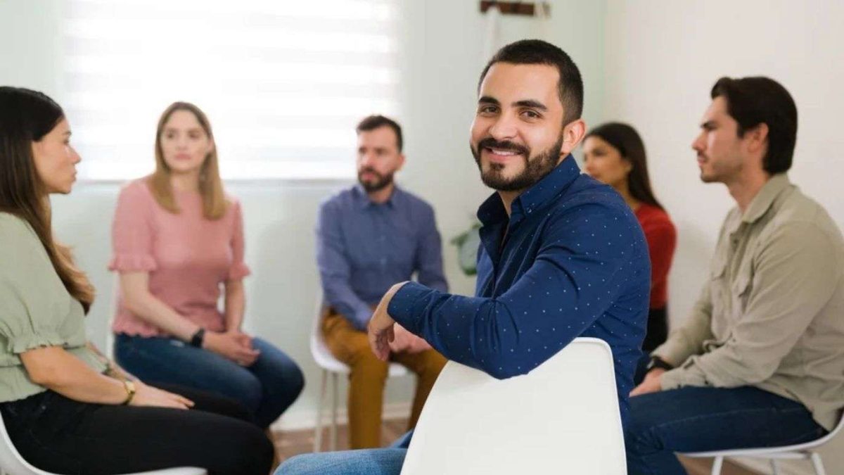 A group of people sitting in a circle during addiction therapy, one man in a blue shirt turning and smiling.