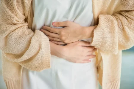A woman wearing a white shirt and beige cardigan holding her two hands across her middle from stomach pain after drinking alcohol.