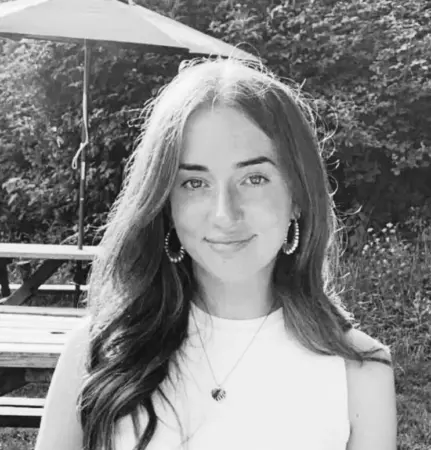 A black and white photo of Katy M smiling in a garden sitting in front of a park bench with a large umbrella.